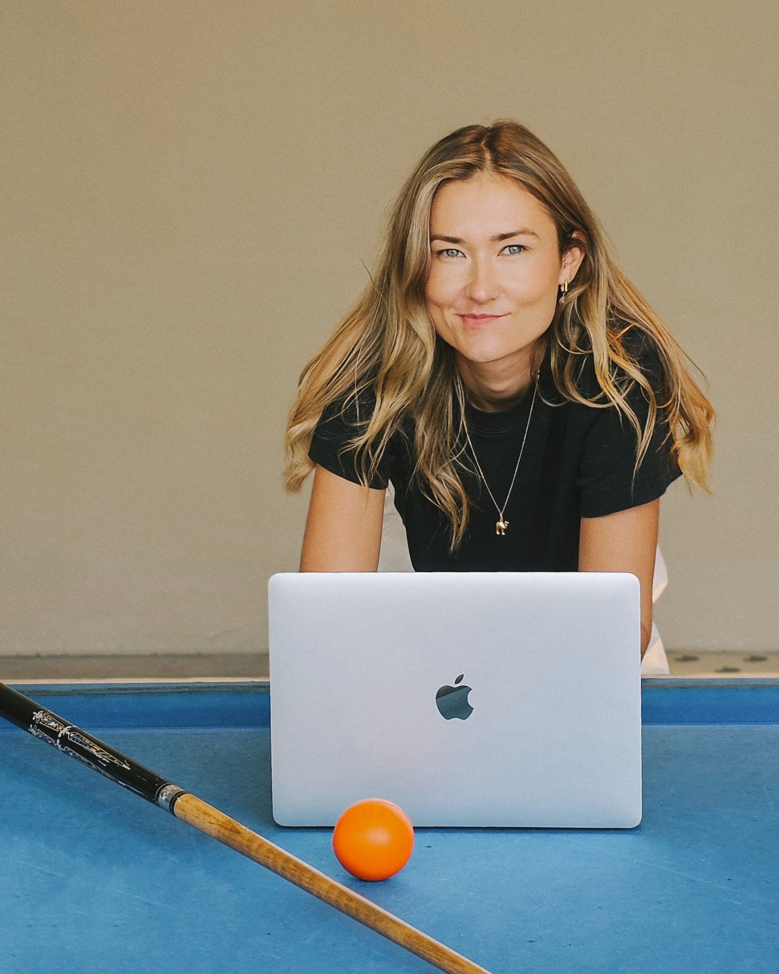 Sintija, founder of June19, leaning on a laptop set on a blue pool table, with an orange ball in front and a pool cue beside her