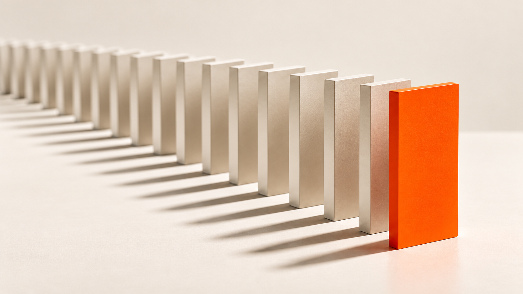 A line of pale off-white domino blocks standing on a cream surface, with the block closest to the camera painted bright orange and every block casting a long shadow